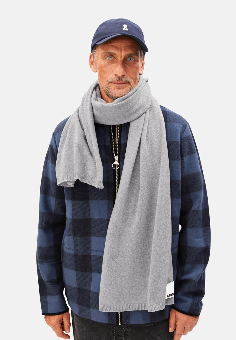 Écharpe en laine grise drapée autour du cou, portée avec une veste à carreaux bleu marine, dotée d'un zip à l'avant et de détails de poches.