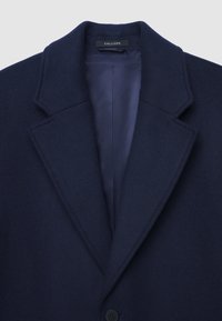 Cappotto in lana blu navy scuro con rever, foderato in modo liscio e un singolo bottone visibile, etichettato "CALLIOPE" all'interno del colletto.