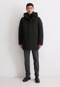 Schwarze Parka mit Kapuze, ausgestattet mit einem Frontreißverschluss und Taschen. Tragen Sie sie über einem schwarzen Pullover und grauen Hosen, kombiniert mit schwarzen Schuhen.