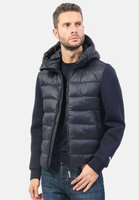 Gilet imbottito blu navy con cappuccio, chiusura con zip, texture trapuntata prominente e tasche laterali, indossato sopra un maglione scuro.