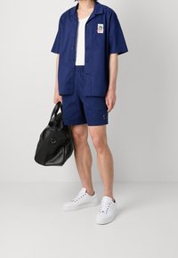 Mörkblå kortärmad skjorta och matchande shorts, tillverkade av ett slätt tyg, med en vit linne under och svart väska. Vita sneakers fulländar looken.