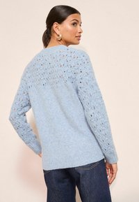 Svetlomodrý pletený sveter s dierkovaným vzorom na ramenách a dlhými rukávmi. Rebrovaný lem, mäkká textura, kombinovaný s tmavými džínami.