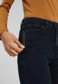 Person som bär mörkblå jeans och en senapsgul långärmad tröja, håller i och delvis drar ner dragkedjan på jeansens framficka.