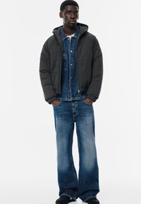 Puffjakke i mørkegrå, lagd over en blå denimskjorte. Løse blå jeans med en slidt finish, båret med sorte fodtøj.
