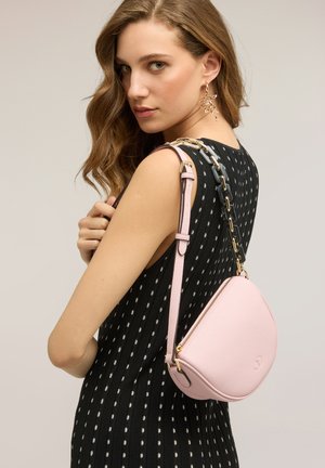 Kvinde med bølget hår iført en sort polkaprikket kjole og guldbelagte øreringe med blade, bærer en lyserød lædertaske med kæderem over skulderen.