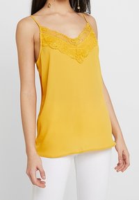 Camisole jaune avec des bordures en dentelle au niveau du décolleté, fines bretelles et une texture de tissu lisse. Associé à un pantalon blanc.