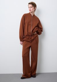 Giacca marrone con cerniera e collo alto, abbinata a pantaloni larghi coordinati. Tessuto morbido con dettagli di texture minimi. Vestibilità casual.