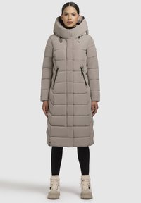 Woman standing wearing a long beige quilted puffer coat with a high hood, black leggings, and beige lace-up boots.