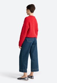Suéter de punto rojo de peluche, ajustado de manera holgada, combinado con jeans de mezclilla de tiro alto y perneras anchas en azul. Llevado con zapatos planos metálicos.