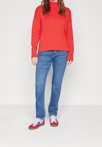 Červený pletený svetr s vysokým límcem, modré rovné džíny a vícebarevné tenisky (červené, růžové, modré) s bílými tkaničkami a hnědými podrážkami.