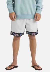 Man wearing light gray shorts with floral trim, a light blue sweatshirt, and brown flip-flops standing against a white background.