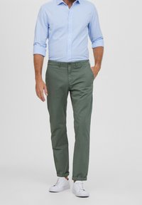 Zelené bavlněné chinos kalhoty s bočními kapsami, ohrnutým lemem a hnědým knoflíkem v pase, doplněné světle modrou košilí s dlouhým rukávem a bílými teniskami.