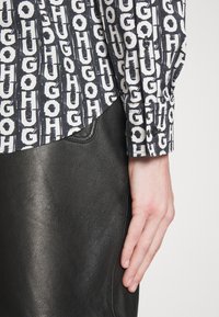 Main reposant à côté d'une jupe en cuir noire, portant une chemise à manches longues noir et blanc avec un motif de texte en gras répété "HUGO".