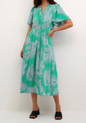 Femme portant une robe midi verte avec un motif paisley violet clair et des manches flottantes, associée à des sandales noires à talon bloc ouvertes à l'avant.
