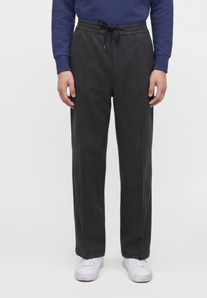 Personne portant un pantalon gris foncé à cordon, des baskets blanches et un haut bleu à manches longues, debout devant un fond uni.