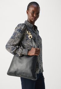 Black leather shoulder bag with a smooth texture, gold hardware accents, and a triangular shape. Model wears a dotted blouse.