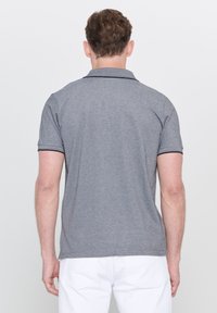 Hombre de espaldas vestido con una camiseta de polo gris de manga corta y pantalones blancos, frente a un fondo claro y liso.