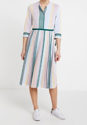 Vrouw draagt een knielange, lange mouwen, gestreepte jurk in pasteltinten met witte sneakers, staand tegen een effen witte achtergrond.