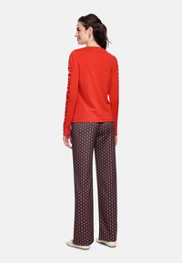 Red long-sleeve top with gathered detail on sleeves, paired with wide-leg patterned trousers featuring a diamond design in brown and cream.