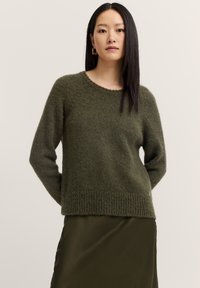 Dunkelgrüner gestrickter Pullover mit geripptem Saum und Bündchen, mit rundem Halsausschnitt und weicher Textur, getragen über einem satinierenden dunkelgrünen Rock.