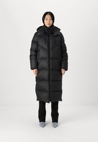 Woman wearing a long black puffer coat with hood, black pants, and light blue pointed-toe shoes, standing against a white background.