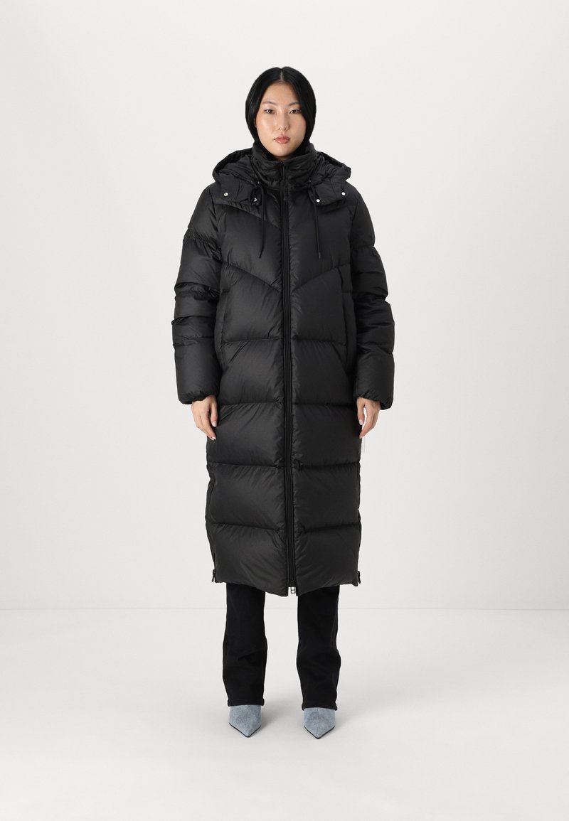 Woman wearing a long black puffer coat with hood, black pants, and light blue pointed-toe shoes, standing against a white background.