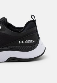 Svart atletisk sko med nät och syntetiskt material, med en vit Under Armour-logotyp och en dämpad sula. Släta och texturerade accenter.