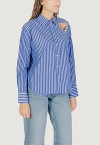 Camicia a maniche lunghe a righe blu e bianche con un ornamento a forma di cuore sul petto. Tessuto in cotone con una consistenza morbida.