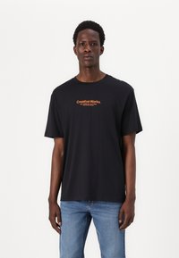 Sort bomulds t-shirt med korte ærmer, med orange tekst "Creative Works." på brystet. Lige skåret design, standard pasform.