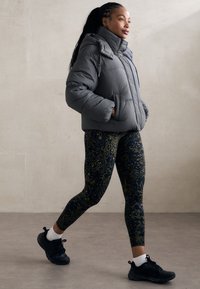 Žena kráčející uvnitř v šedé péřové bundě, vzorovaných legínách, bílých ponožkách a černých sportovních botách.