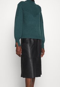 Pull en maille vert profond avec un empiècement texturé, col montant et manches larges, associé à une jupe en cuir noire mi-longue avec des coutures en panneaux.