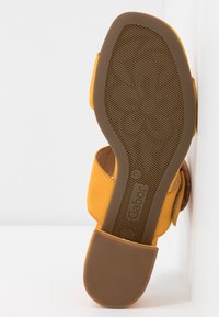 Sandale en daim jaune avec de larges brides et une semelle en caoutchouc marron, dotée d'une texture antidérapante et d'un logo de marque embossé sur la semelle.