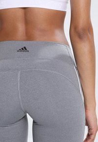 Detailní záběr osoby ve šedých legínách Adidas a bílém sportovním podprsenkovém topu Adidas, zdůrazňující loga a texturu tkaniny.