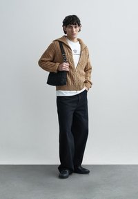 Suéter marrón de punto con cremallera frontal, combinado con una camisa blanca y pantalones negros de pierna ancha. Bolso negro acolchado y zapatos negros.
