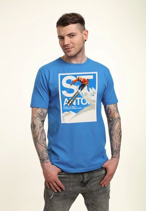 Jonge man met kort haar en tatoeages, gekleed in een blauw T-shirt met een skiër op een besneeuwde berg en de tekst "St. Anton".