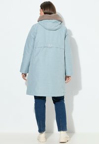 Cappotto lungo celeste chiaro con colletto in peluche beige, tasche laterali e dettaglio del logo sul retro, indossato con jeans blu e stivaletti beige.