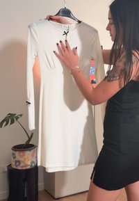 Vestido branco de malha com mangas compridas e decote redondo, apresentando um pequeno detalhe em laço preto na frente e silhueta ajustada.