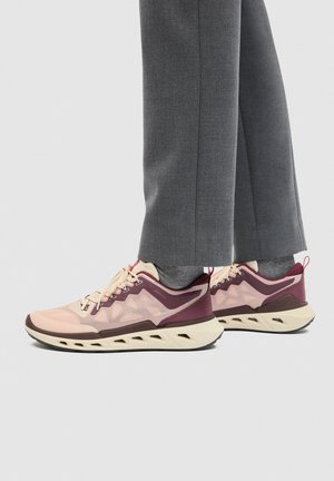 Personne portant un pantalon gris et des chaussures de sport beige et bordeaux à semelles épaisses debout sur un sol gris clair.
