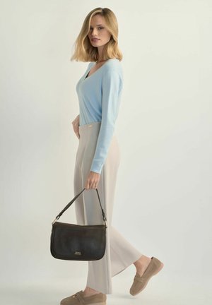 Kobieta o blond włosach ubrana w jasnoniebieski sweter i beżowe spodnie, trzymająca czarną torebkę, stoi na tle jednolitego jasnego tła.