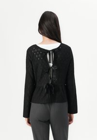 Top in pizzo nero con maniche lunghe e design peplum. La parte posteriore presenta un taglio a chiave fissato con nastri neri. Indossato sopra pantaloni grigi.
