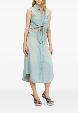 Donna con vestito in denim chiaro senza maniche con laccio e bottoni sul davanti, abbinato a scarpe in denim con tacco alto e punta aperta con dettagli di frange.