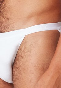 Close-up van de heup en bovenbeen van een man die witte ondergoed draagt, met zichtbare lichaamshaar en huidtextuur.
