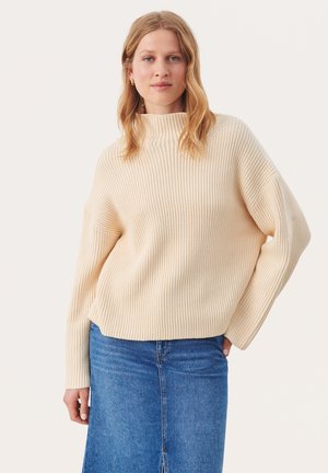 Pull en maille côtelée crème avec col haut et épaules tombantes, associé à une jupe en denim, affichant une silhouette décontractée et oversized.