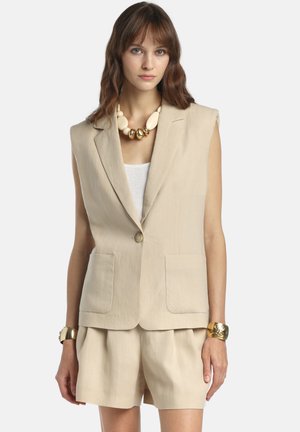 Žena oblečená v béžovom bezrukávovom blejzri a zodpovedajúcich kraťasoch, doplnená o masívny náhrdelník a zlaté náramky, na jednoduchom pozadí.