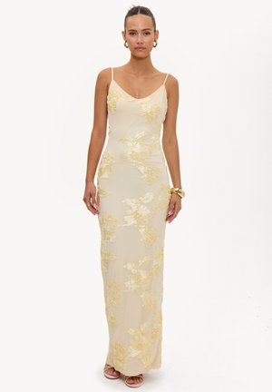 Femme portant une robe longue crème ajustée avec une broderie florale jaune, des bretelles spaghetti, des boucles d’oreilles créoles dorées et un bracelet en or.