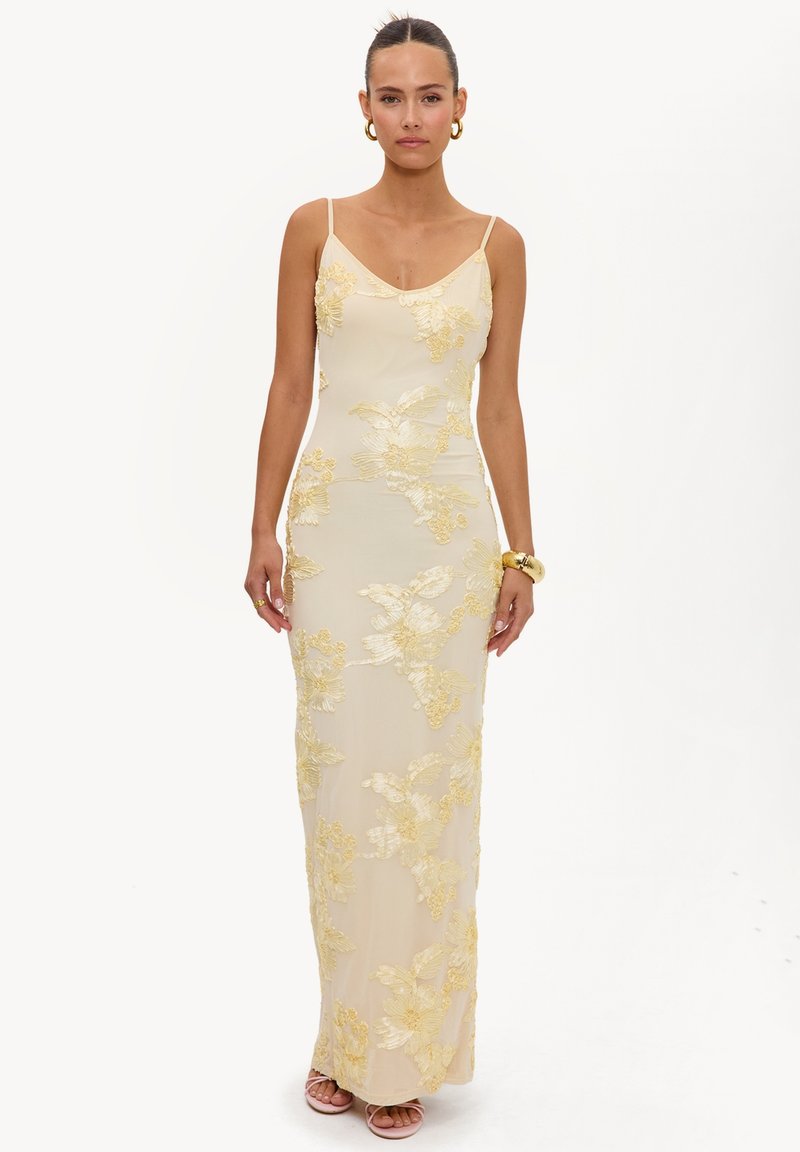 Femme portant une robe longue crème ajustée avec une broderie florale jaune, des bretelles spaghetti, des boucles d’oreilles créoles dorées et un bracelet en or.