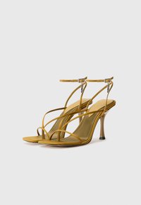Sandales à talons hauts jaunes avec des lanières, une texture douce et lisse, des brides fines autour de la cheville et un bout pointu. Le talon transparent ajoute une touche moderne.