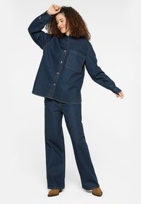 Dark denim oversized shirt with button front, single chest pocket, and long sleeves, paired with wide-leg dark denim pants. Brown boots.