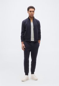 Άνδρας που φοράει μπλε σκούρο μπουφάν με φερμουάρ και παντελόνι, γκρι μπλουζάκι και λευκά αθλητικά παπούτσια, στέκεται με το ένα χέρι στην τσέπη σε απλό λευκό φόντο.