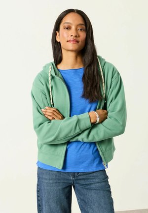 Ung kvinde med langt mørkt hår iført en grøn hættetrøje med lynlås over en blå t-shirt og blå jeans, stående med armene krydsede.